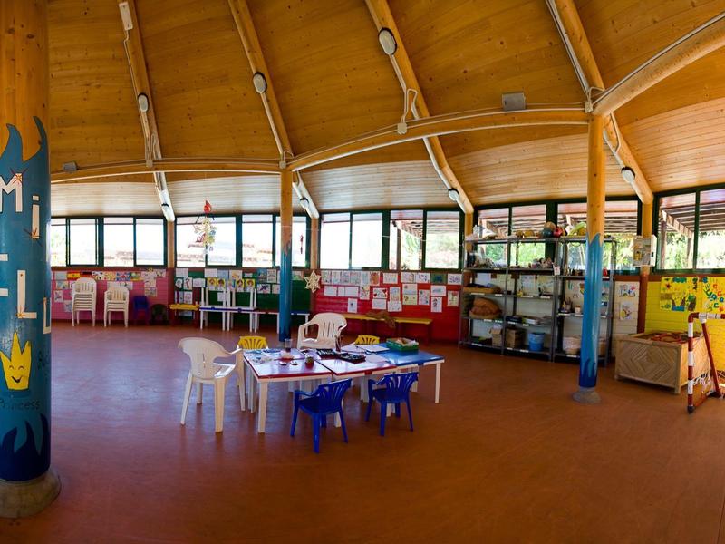 Großer, heller Raum mit Holzdach, bunten Stühlen, Tischen und Säulen mit Kindermalereien.