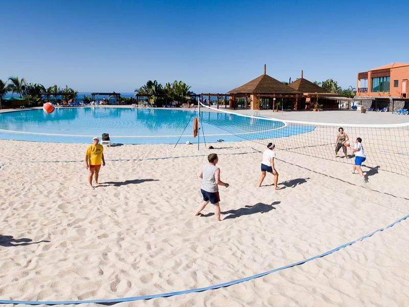 Menschen spielen Volleyball auf großem Sandstrand vor großem blauem Pool und Gebäuden bei klarem Himmel.