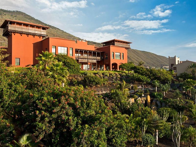 Großes rotes Haus auf Hügel mit vielen grünen Bäumen und blauem Himmel im Hintergrund.