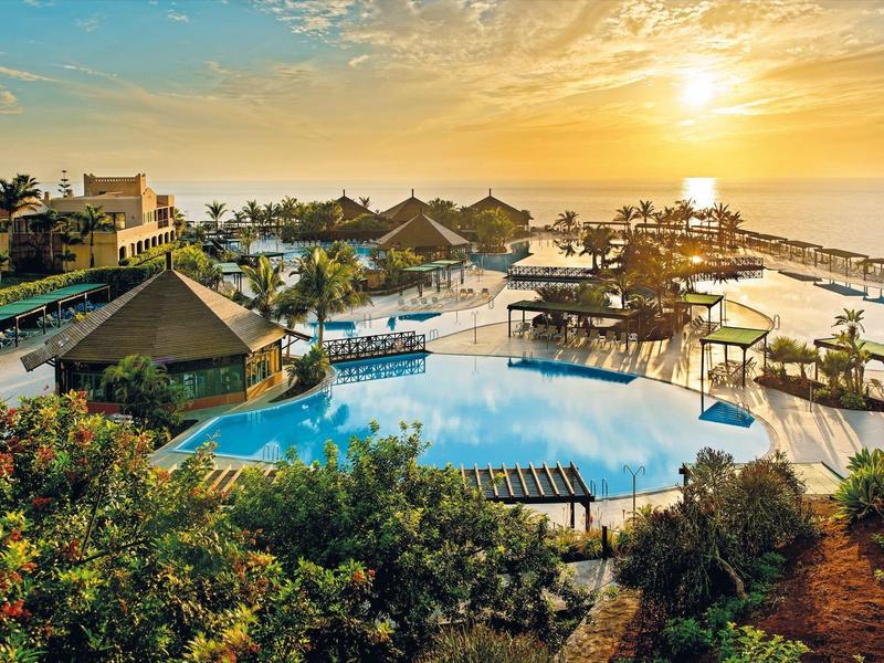 Ein großer Pool mit Pavillons am Strand bei Sonnenuntergang, umgeben von Palmen und Gebäuden.