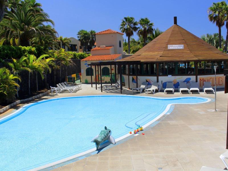 Außerhalb mit klarem blauen Pool, Liegestühlen, Palmen und Gebäuden unter blauem Himmel.