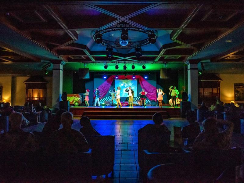 Bühne mit bunten Lichtern und Darstellern vor einem Publikum in einem großen Theatersaal.