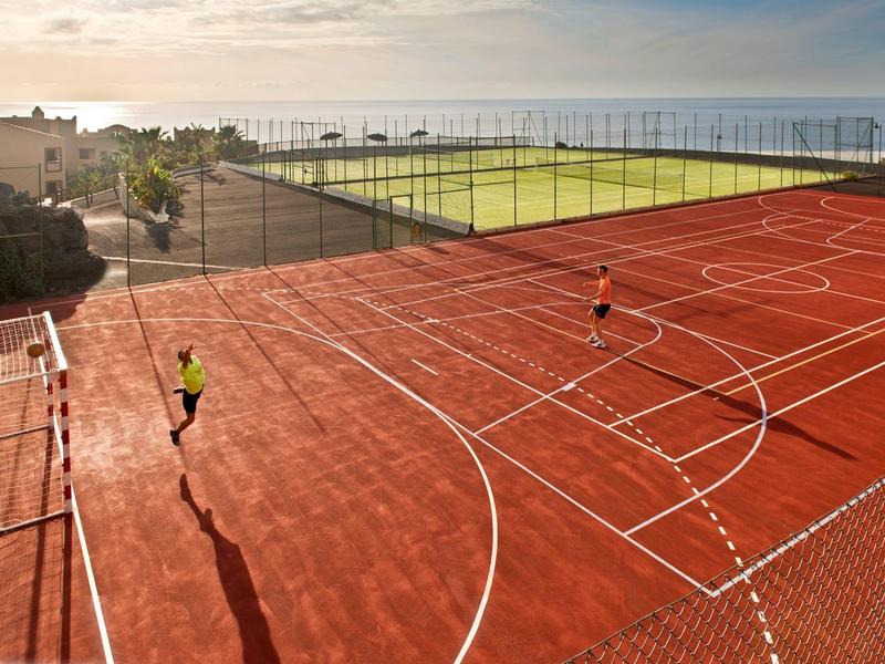 Sportplatz mit rotem Boden, zwei Personen spielen Basketball, Blick aufs Meer im Hintergrund.