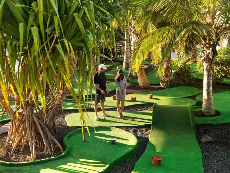 Minigolfplatz mit grünen Teppichen, Palmen und zwei Personen beim Spielen.