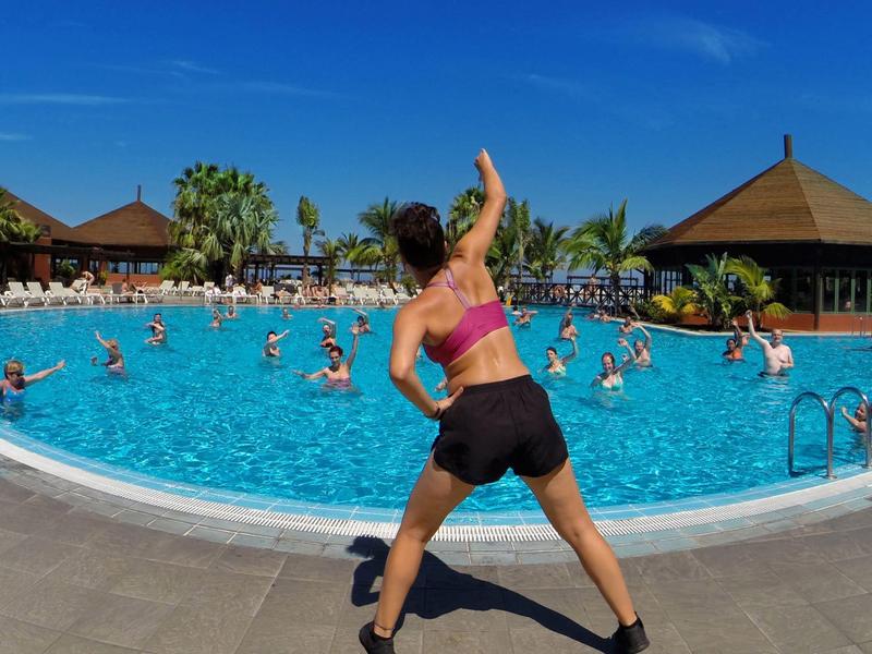 Frau macht Gymnastik vor großem Pool mit vielen Menschen und Holzhäusern bei sonnigem Himmel.