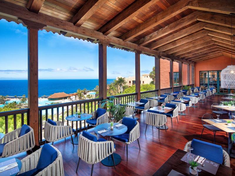 Panorama-Restaurant mit Holzboden, blau-weißen Stühlen und Meerblick bei klarem Himmel.