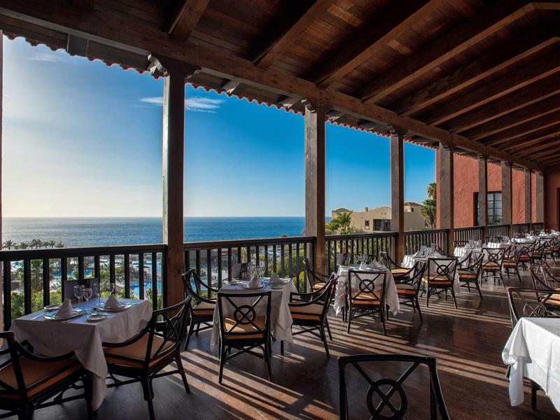 Überdachte Terrasse mit gedeckten Tischen, Holzbalken und Meerblick im Sonnenlicht.