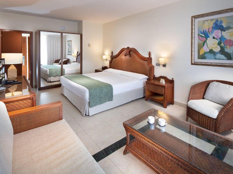 Ein gemütliches Hotelzimmer mit Doppelbett, Sofa, Sessel, Glas-Couchtisch und dekorativem Bild an der Wand.