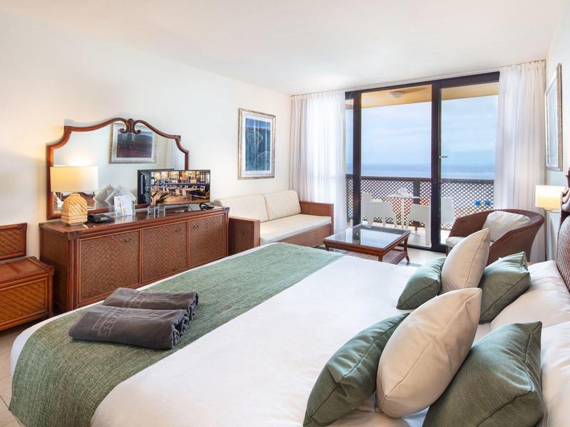 Helles Schlafzimmer mit grünem Bett, Holzmöbeln und großer Fensterfront mit Meerblick.