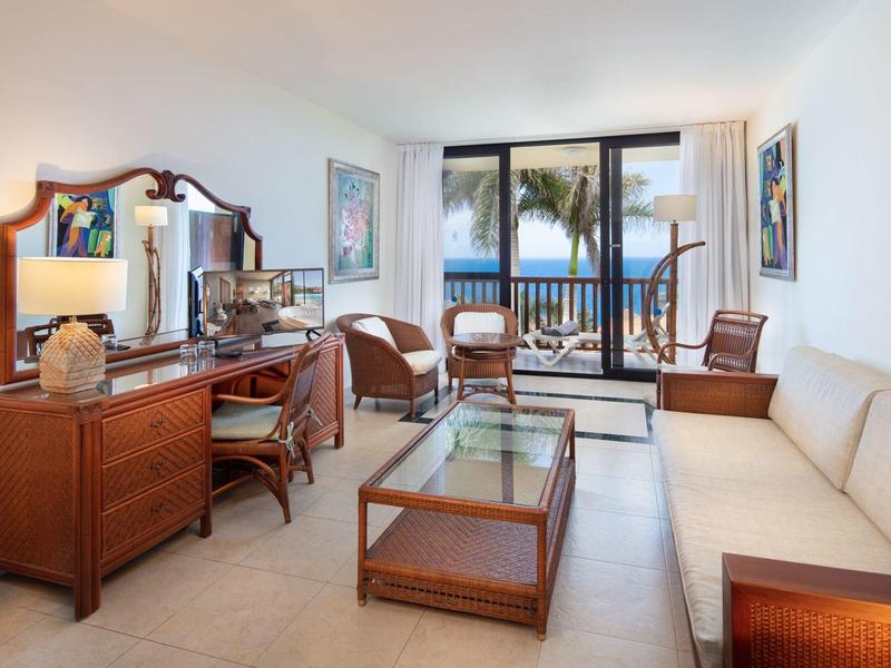 Helles Wohnzimmer mit Holzmöbeln, Glas-Couchtisch und Meeresblick durch große Fenster.