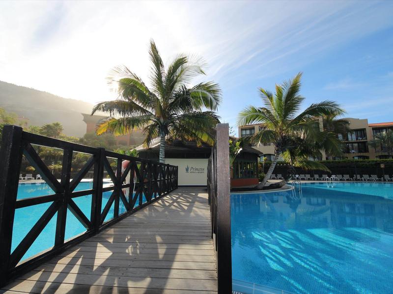 Brücke aus Holz über türkisfarbenes Schwimmbecken mit Palmen und Häusern im Sonnenlicht.