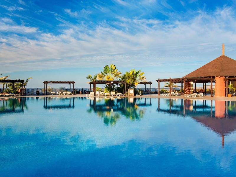 Großer blauer Pool mit klarer Spiegelung, umgeben von Holzhütten und Palmen unter blauem Himmel.