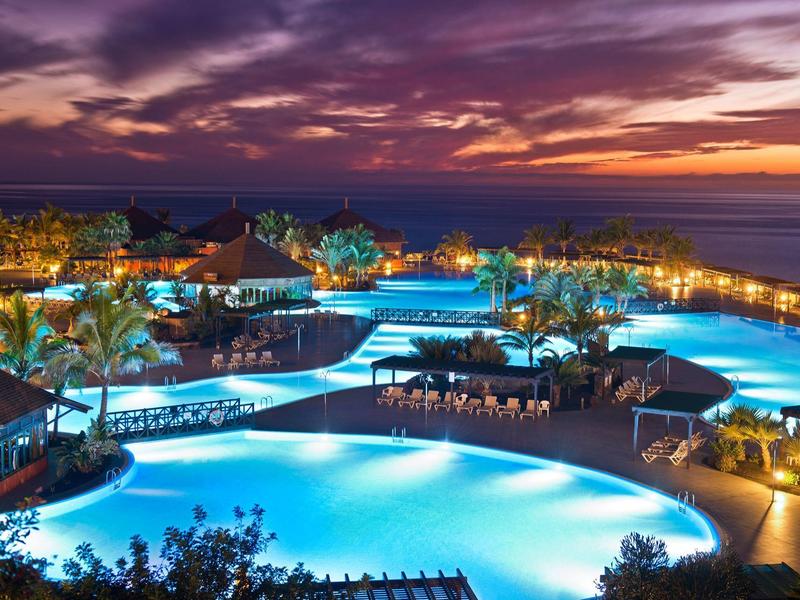 Leuchtender Poolkomplex mit Palmen und Sitzbereichen bei Sonnenuntergang am Meer.