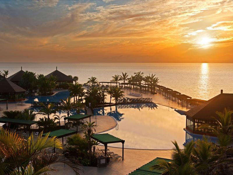 Ein großer Hotelpool mit Liegestühlen, Palmen und Sonnenuntergang über ruhigem Meer.