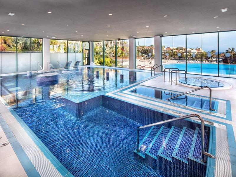 Modernes Hallenbad mit großen Glasfenstern, blauen Fliesen und verschiedenen Schwimmbecken.