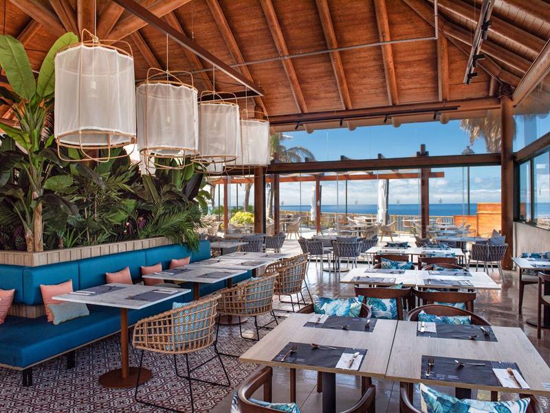 Modernes Restaurant mit blauen Polstersesseln, weißen Tischen, grünen Pflanzen und Meeresblick.