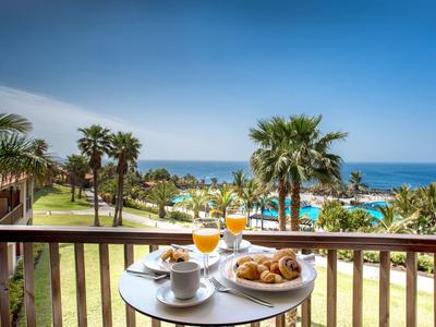 Balkontafel met ontbijt met uitzicht op zee en palmbomen onder een blauwe lucht.