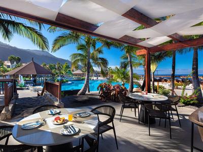 Openlucht restaurant met uitzicht op zwembad, palmbomen en bergen op een zonnige dag.