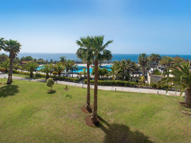 Uitzicht over een groen gazon met palmbomen en zwembad tot aan zee bij een heldere lucht.