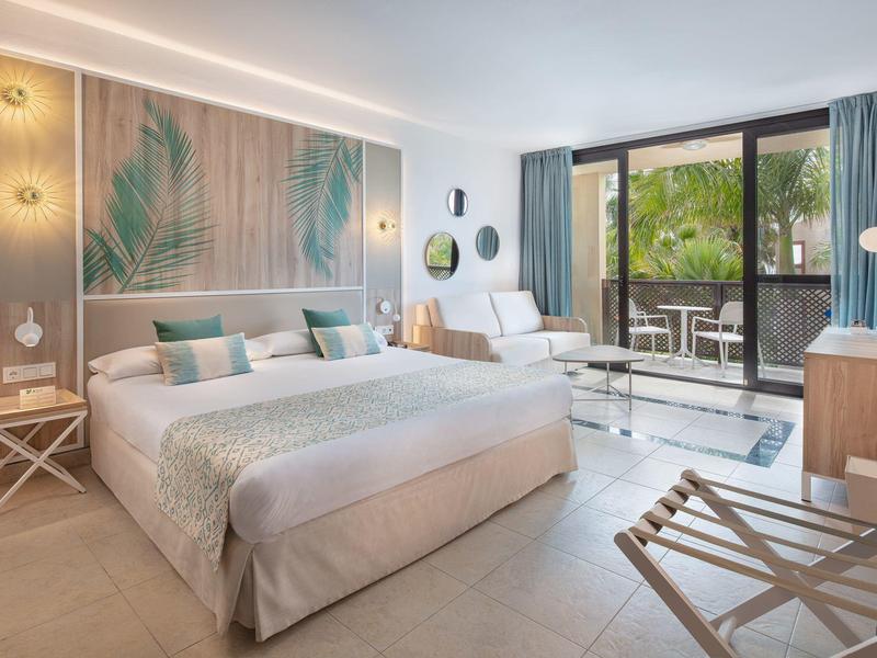 Lichte hotelkamer met groot bed, zithoek en balkon met uitzicht op groen.