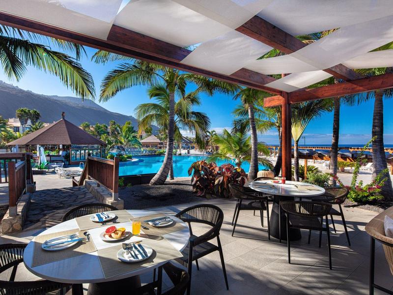 Openlucht restaurant met uitzicht op zwembad, palmbomen en bergen op een zonnige dag.