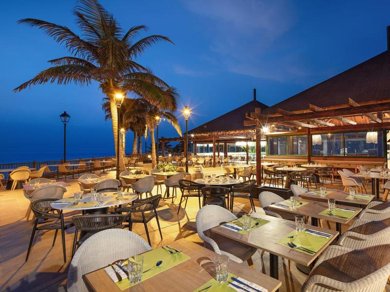 Avondzicht op een terras met stoelen en tafels langs een kust met palmbomen.