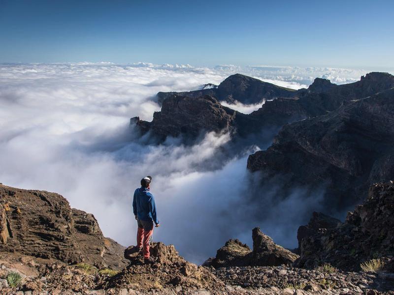 Osoba stoi na skalistym szczycie góry, patrząc na mgłę w dolinie z pasmami gór w tle.
