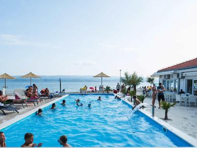 Hotelpool mit Sonnenliegen und Gästen, Meer und klarem Himmel im Hintergrund.