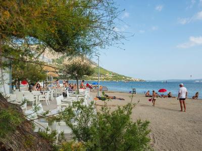 Strand mit vielen Menschen, weißen Stühlen, grünen Bäumen und blauem Himmel am Meer.