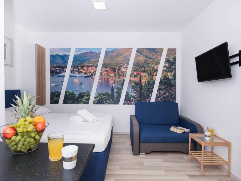 Modern hotel room with bed, sofa, cityscape wall art, TV, and a bowl of fruit.