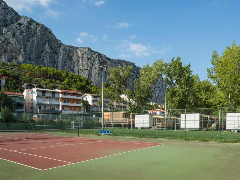 Empty tennis court with mountain scenery and buildings in the background under clear sky.