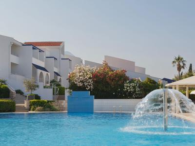 Großer Pool mit Wasserspielen vor modernen weißen Ferienwohnungen bei klarem Himmel.
