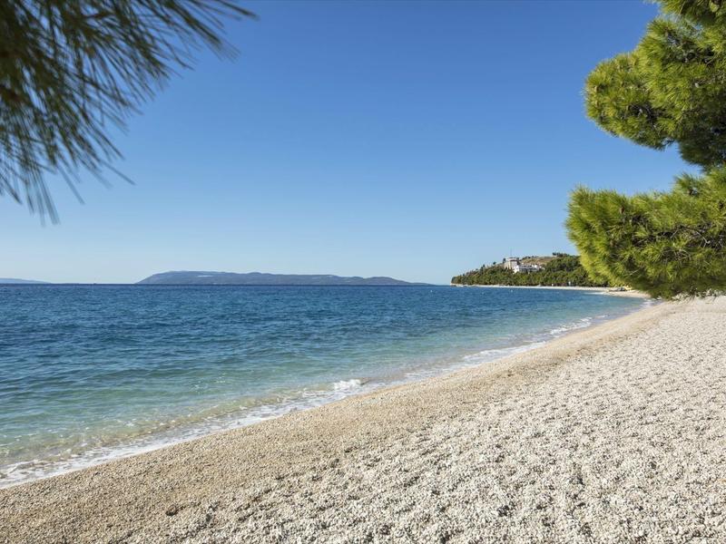 Kieselstrand mit klarem Wasser und blauem Himmel, grüne Bäume am Strand, Insel im Hintergrund.