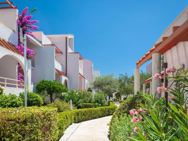 Weg durch gepflegten Hotelgarten mit weißen Gebäuden und blauem Himmel.