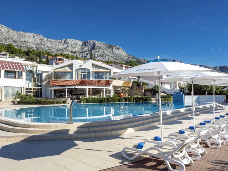 Moderner Außenpool mit Liegestühlen und Sonnenschirmen vor einem Hotel unter blauem Himmel.