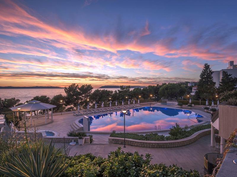 Poolbereich mit Liegestühlen und farbenfrohem Sonnenuntergang am Wasser.