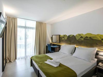 Modernes Hotelzimmer mit Doppelbett, grünem Dekor, Flachbildfernseher und Balkon mit Vorhängen.