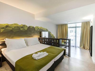 Modernes Hotelzimmer mit großem Bett, grünem Wandbild und Zugang zu einem Balkon mit Vorhängen.