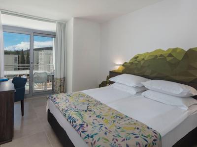 Modernes Hotelzimmer mit Doppelbett, TV, Stuhl und Blick auf eine Terrasse mit Meer.