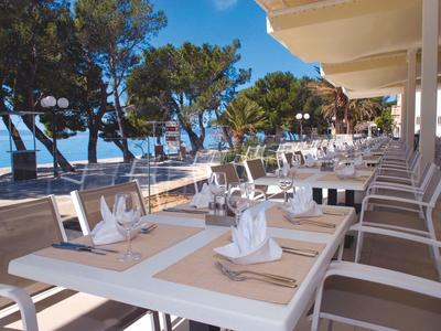 Terrassenrestaurant am Meer mit gedeckten Tischen und Blick auf Pinien bei klarem Himmel.