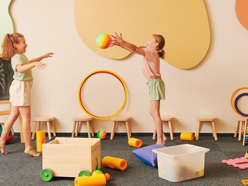 Des enfants jouent avec des jouets colorés dans une pièce lumineuse et confortable avec des chaises en bois et des décorations murales.