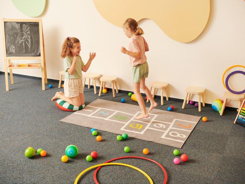 Deux enfants jouent dans une salle de jeux claire avec des jouets et une marelle.