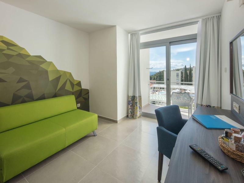 Chambre d'hôtel moderne avec canapé vert, bureau et grande fenêtre donnant sur le port.