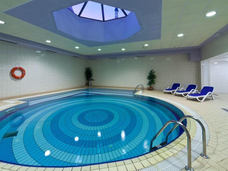 Piscine intérieure ronde avec chaises longues bleues, carreaux blancs et puits de lumière.