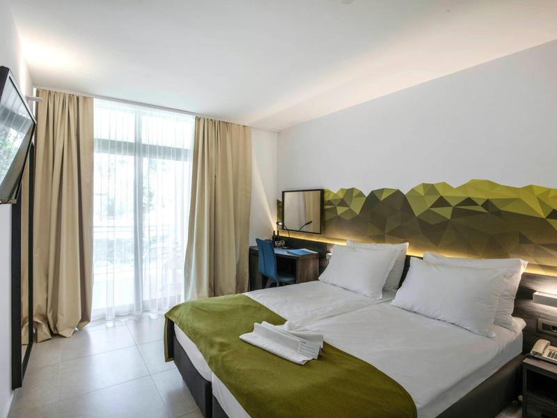 Une chambre d'hôtel moderne avec un lit double, une couverture verte et une fenêtre avec des rideaux beiges.