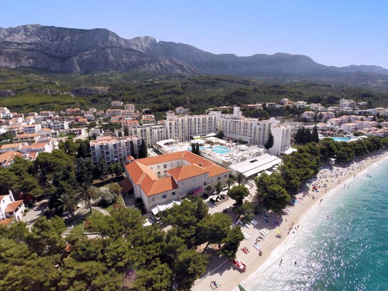 Complexe hôtelier au bord de la mer avec montagnes environnantes et zones boisées par une journée ensoleillée.