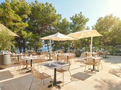 Terrasse eines Café mit Tischen, Stühlen und Sonnenschirmen in einer sonnigen Waldumgebung.