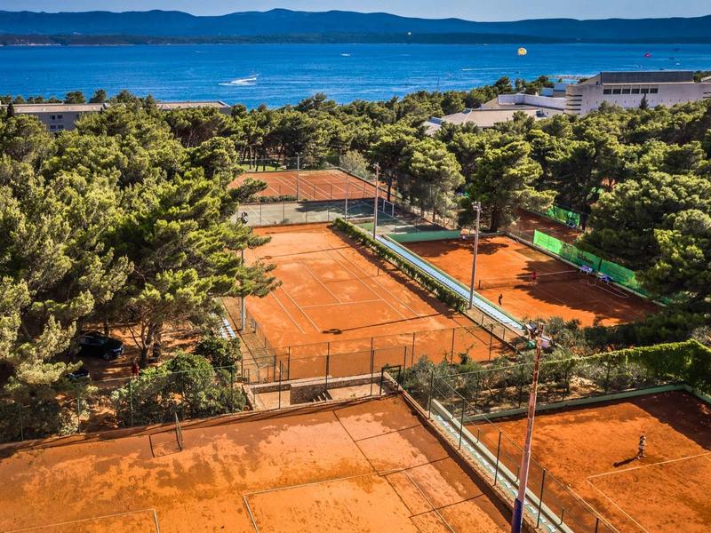 Rote Sand-Tennisplätze umgeben von grünen Bäumen mit Blick auf das blaue Meer und den Himmel.
