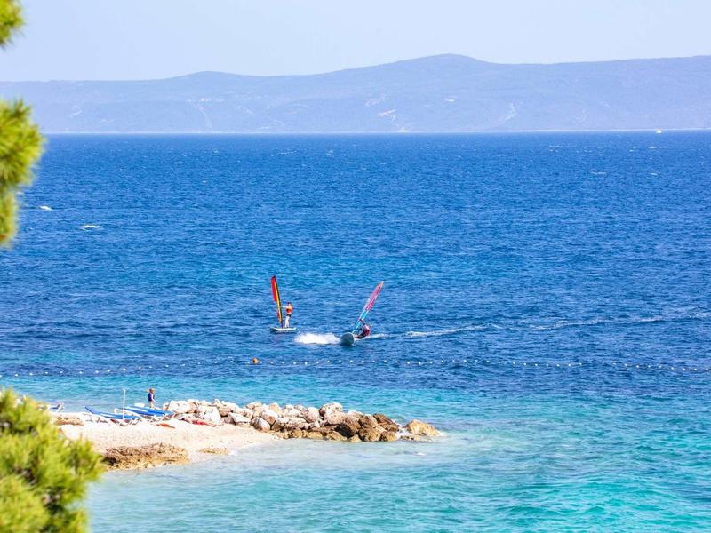 Blaues Meer mit zwei Windsurfern, felsige Küste und grüne Pflanzen im Vordergrund.