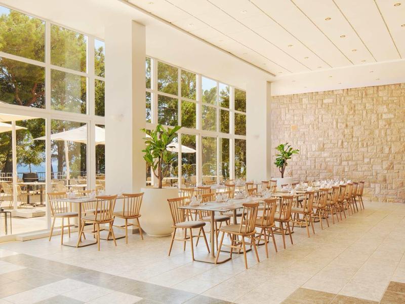 Heller, moderner Speiseraum mit langen Holztischen, Stühlen und großen Fenstern zur Außenterrasse.